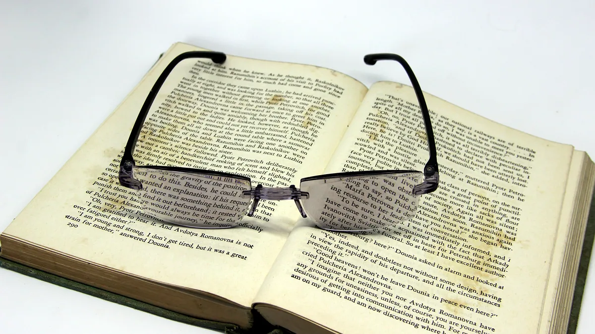 An open book on a wooden desk with a pair of reading glasses laid across the pages.
